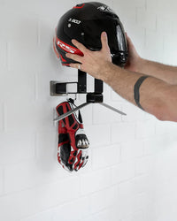Person placing a helmet and gloves on a wall-mounted rack against a white tiled wall.