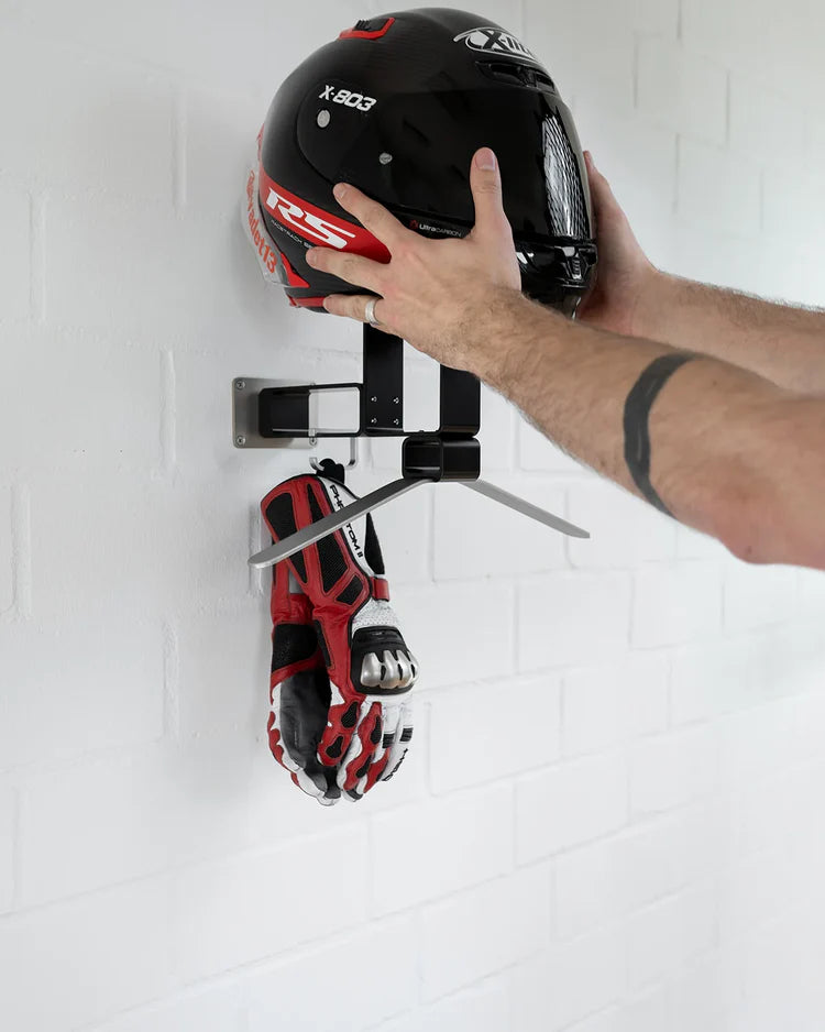 Person placing a helmet and gloves on a wall-mounted rack against a white tiled wall.