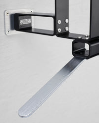Close-up of a black aluminium luggage rack bracket with a brushed silver aluminium support arm, mounted against a light gray wall. The brand name 'SUITBASE' is visible on a small black label.